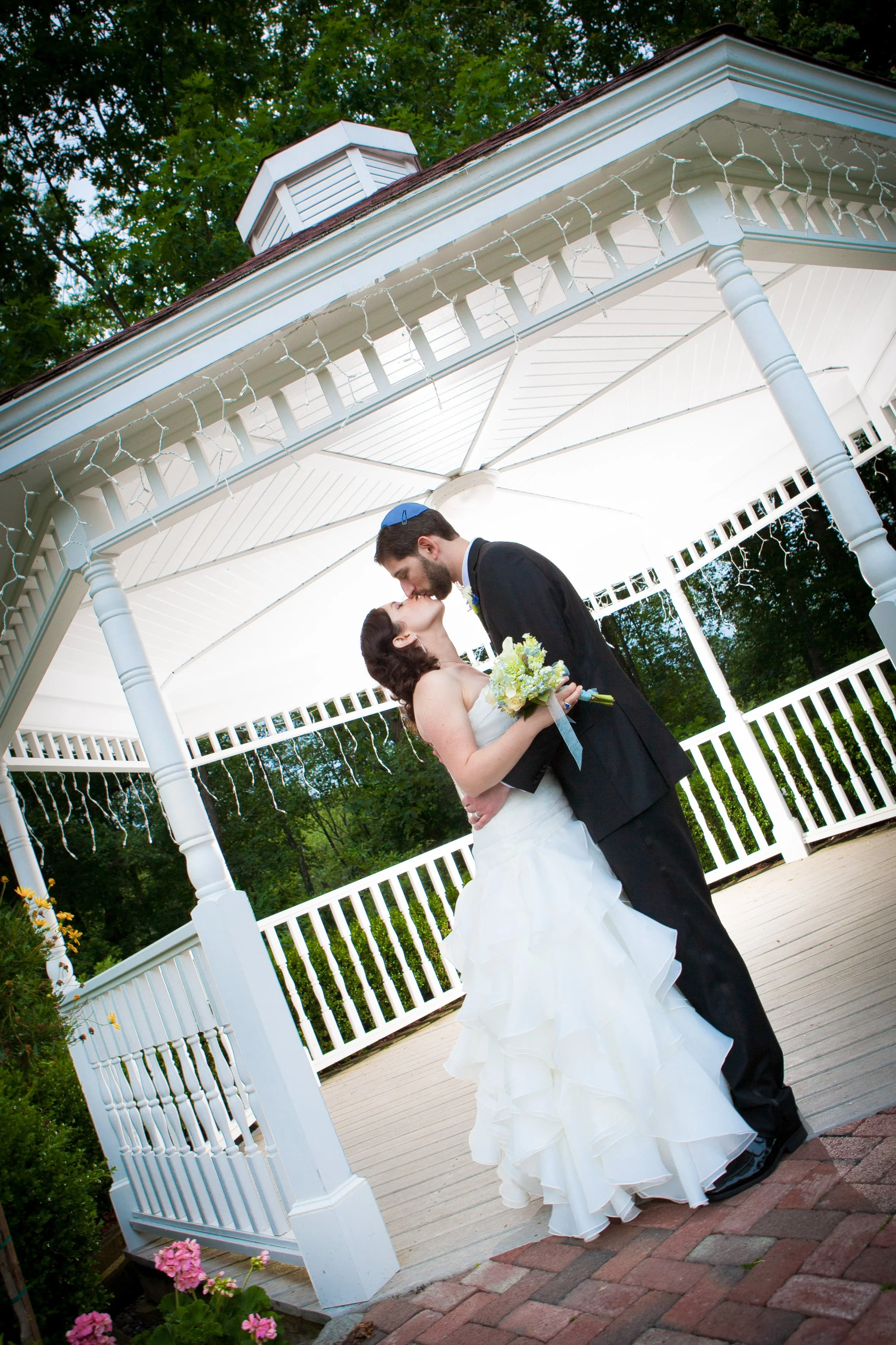 A bride and groom kissing under a white gazebo decorated with string lights, surrounded by greenery and flowers.