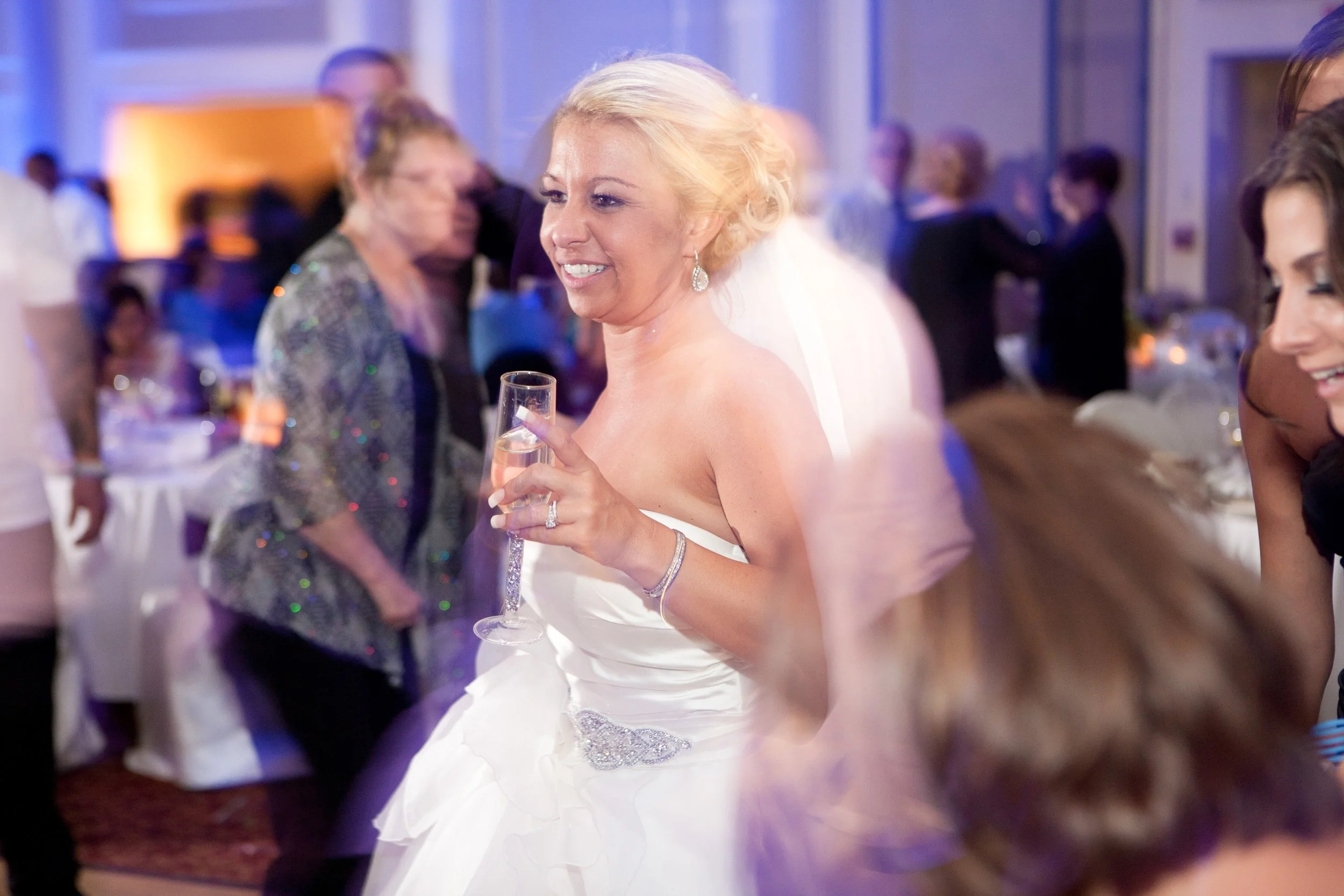 Smiling bride in white wedding dress holding a champagne flute at a wedding reception with guests in background.