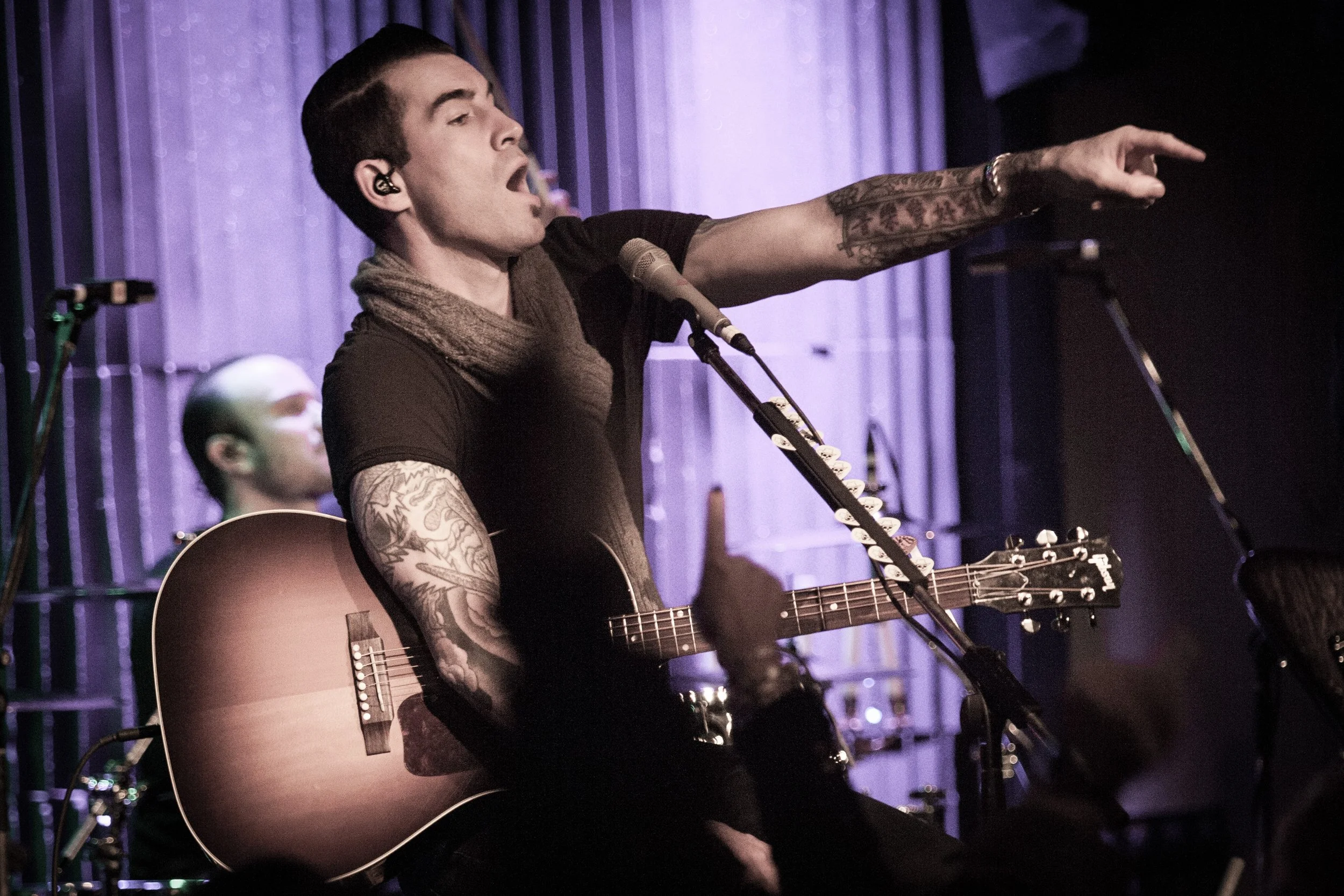 Musician with tattoos performing on stage with an acoustic guitar and microphone