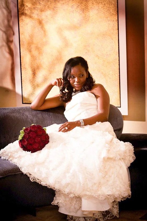 A bride in a wedding dress sitting on a dark couch with a bouquet of red roses, posing with her hand near her face and smiling slightly.