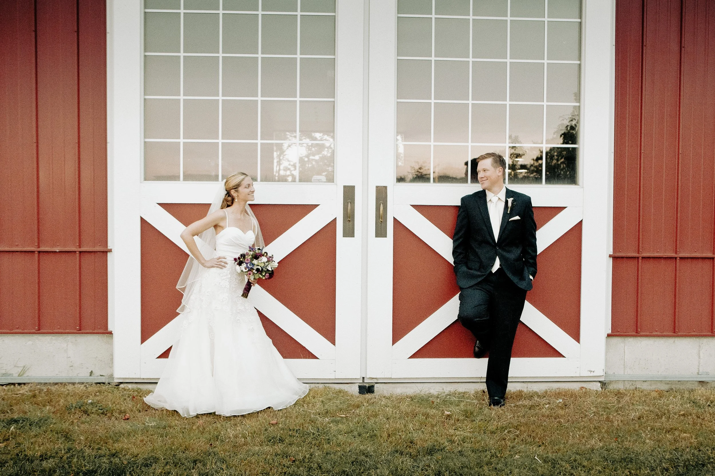 A Colorado wedding bride and groom stand on a grassy area in front of large red and white barn doors, looking at each other with smiles.