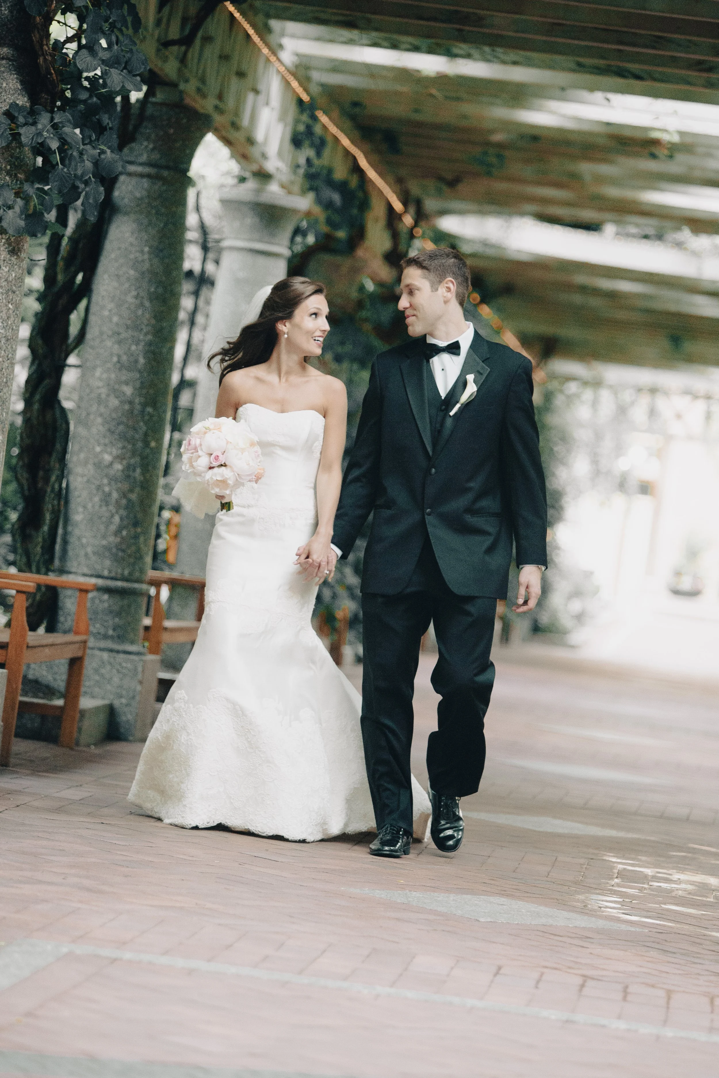 A bride and groom walk hand in hand under a rustic outdoor wooden canopy, smiling and looking at each other during their wedding.