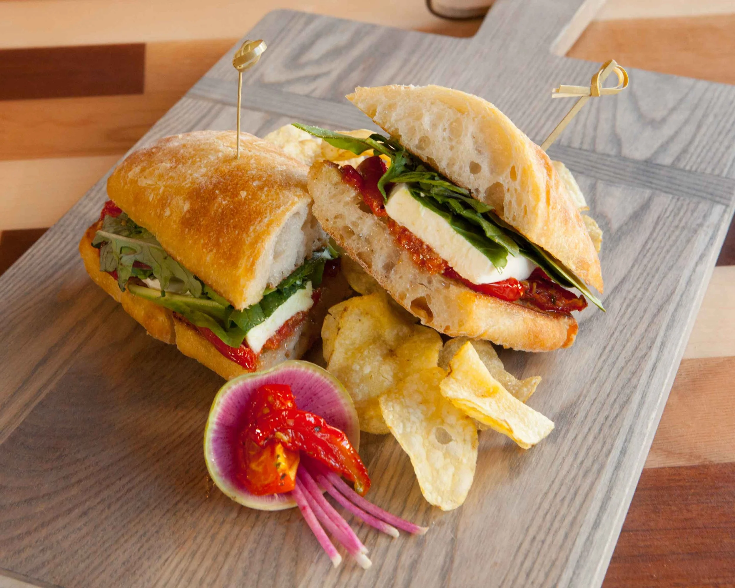 Caprese sandwich with mozzarella, tomatoes, greens, on ciabatta, served with potato chips and pickled vegetables on a wooden board.