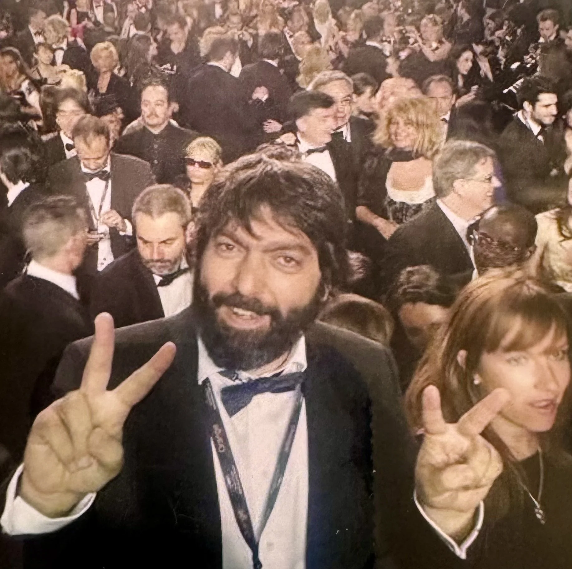 A man with dark hair and a beard, dressed in a tuxedo, making peace signs with both hands at a crowded formal event.