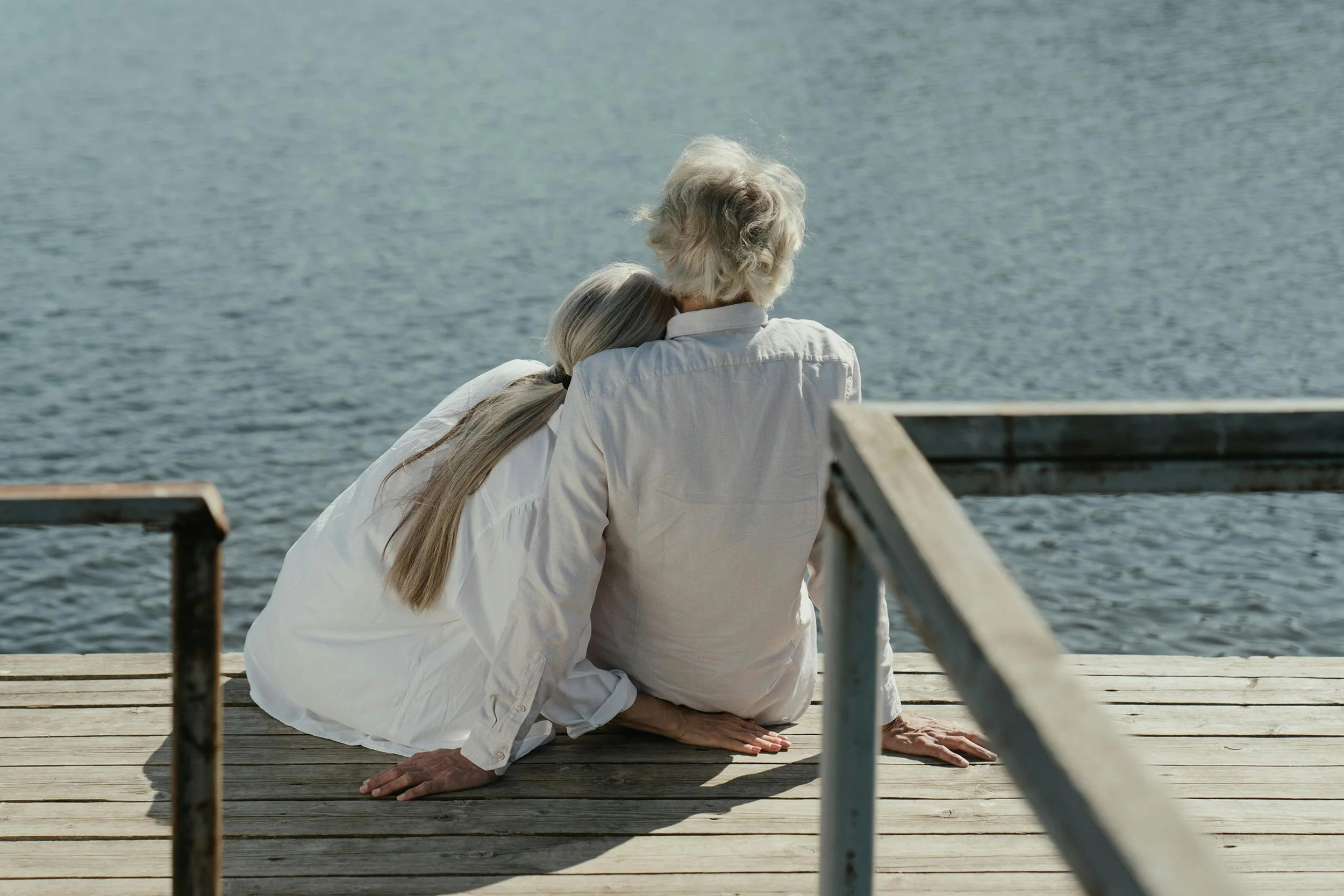 An elderly man and woman sitting on a wooden dock by a body of water, embracing each other while looking out at the water.