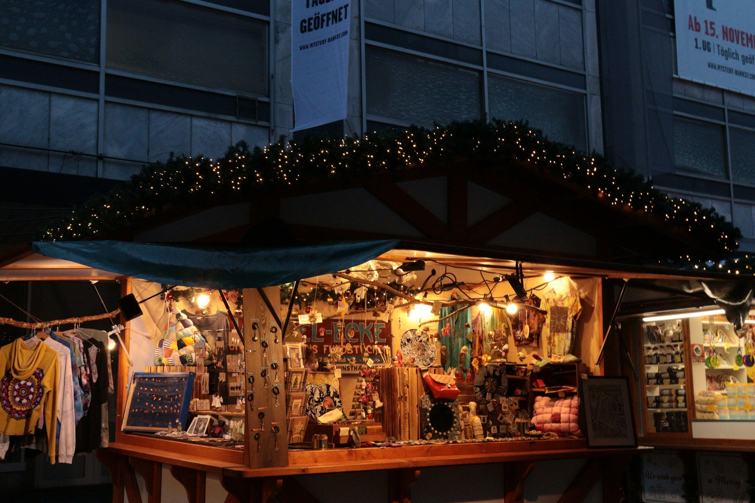 A marketplace stall decorated with Christmas lights, selling various clothing, jewelry, and decorative items at night.