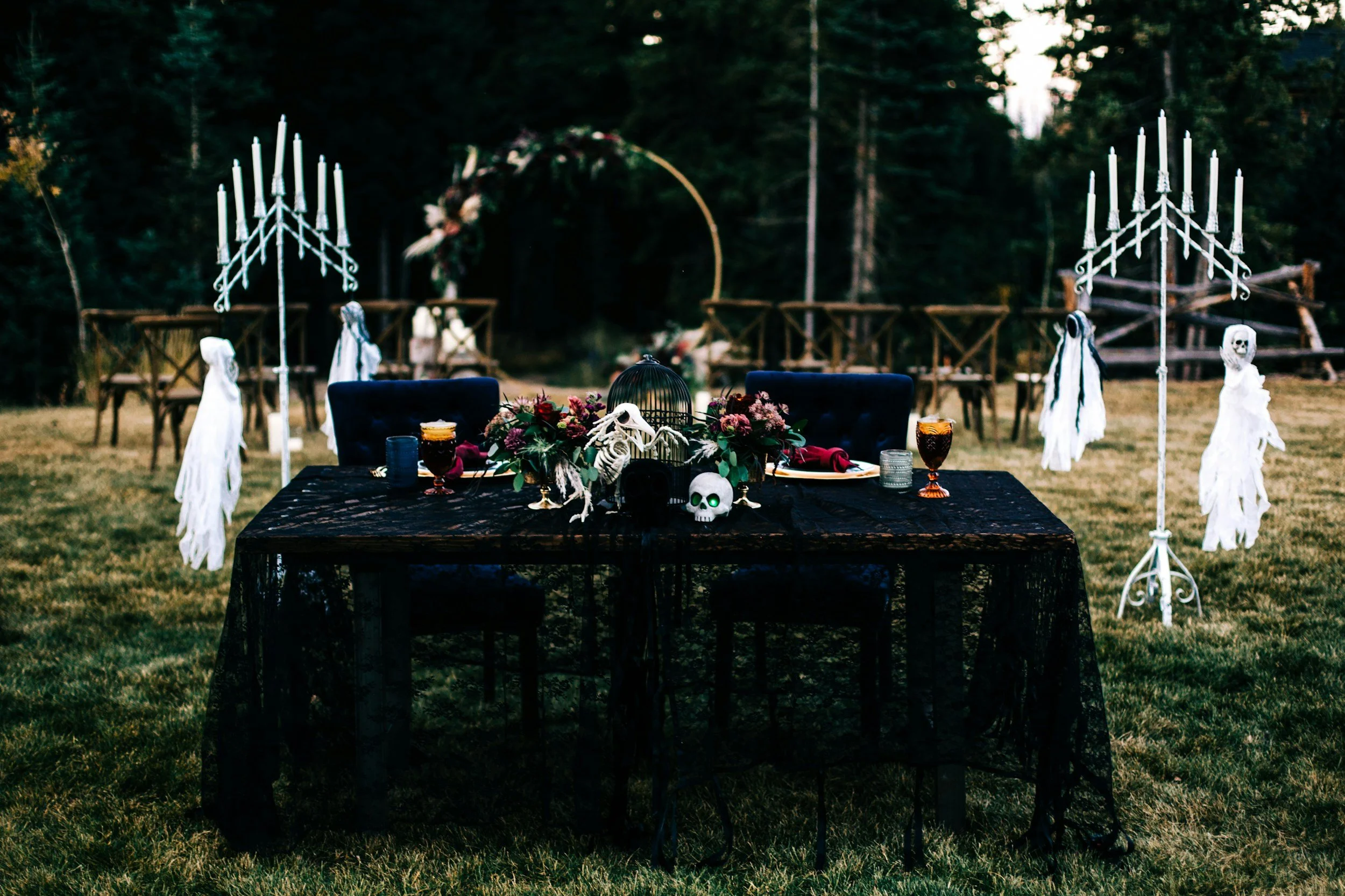 A tablescape with Halloween themed decorations including skulls, skeleton hands, skull glasses, and flowers, set outdoors with candelabra and chairs in the background.