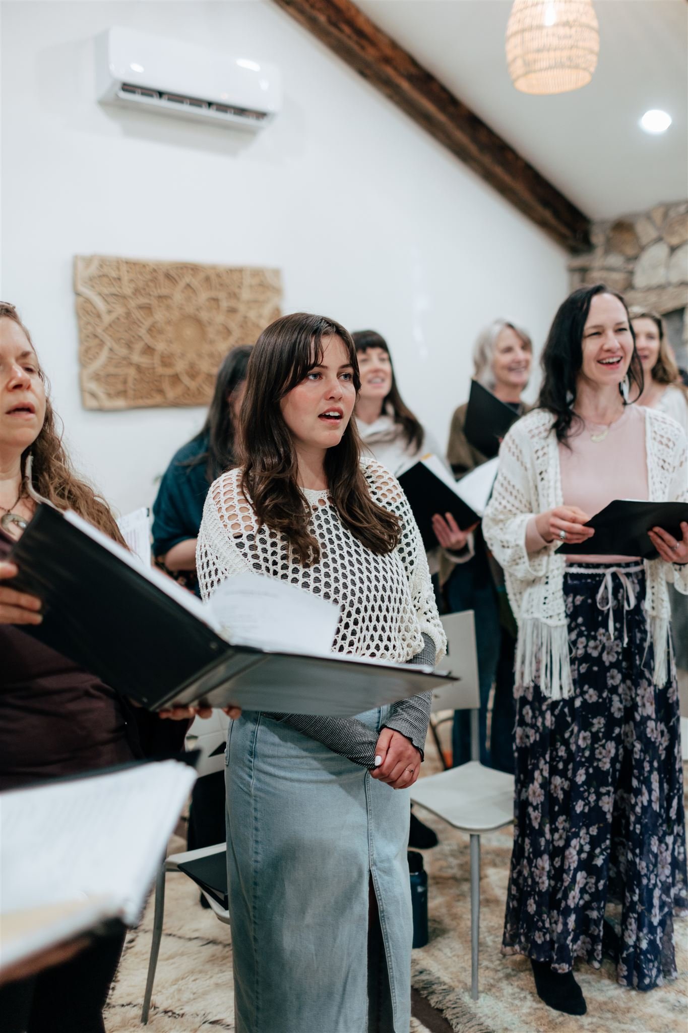 Group of women singing together indoors, holding music sheets.
