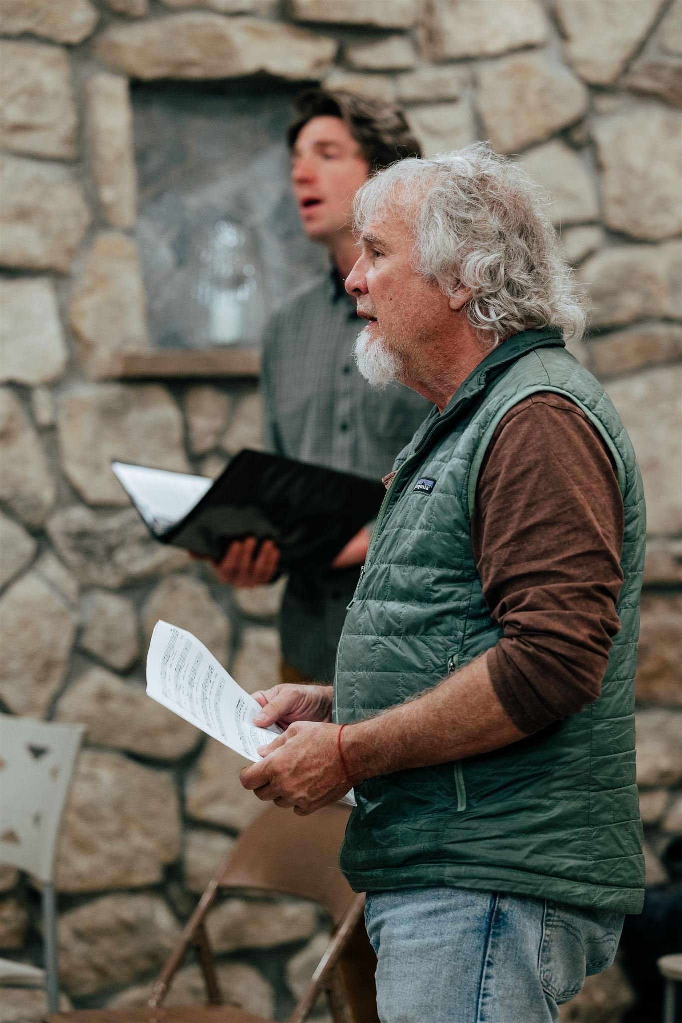 Two men singing together indoors, one holding sheet music, in front of a stone wall.