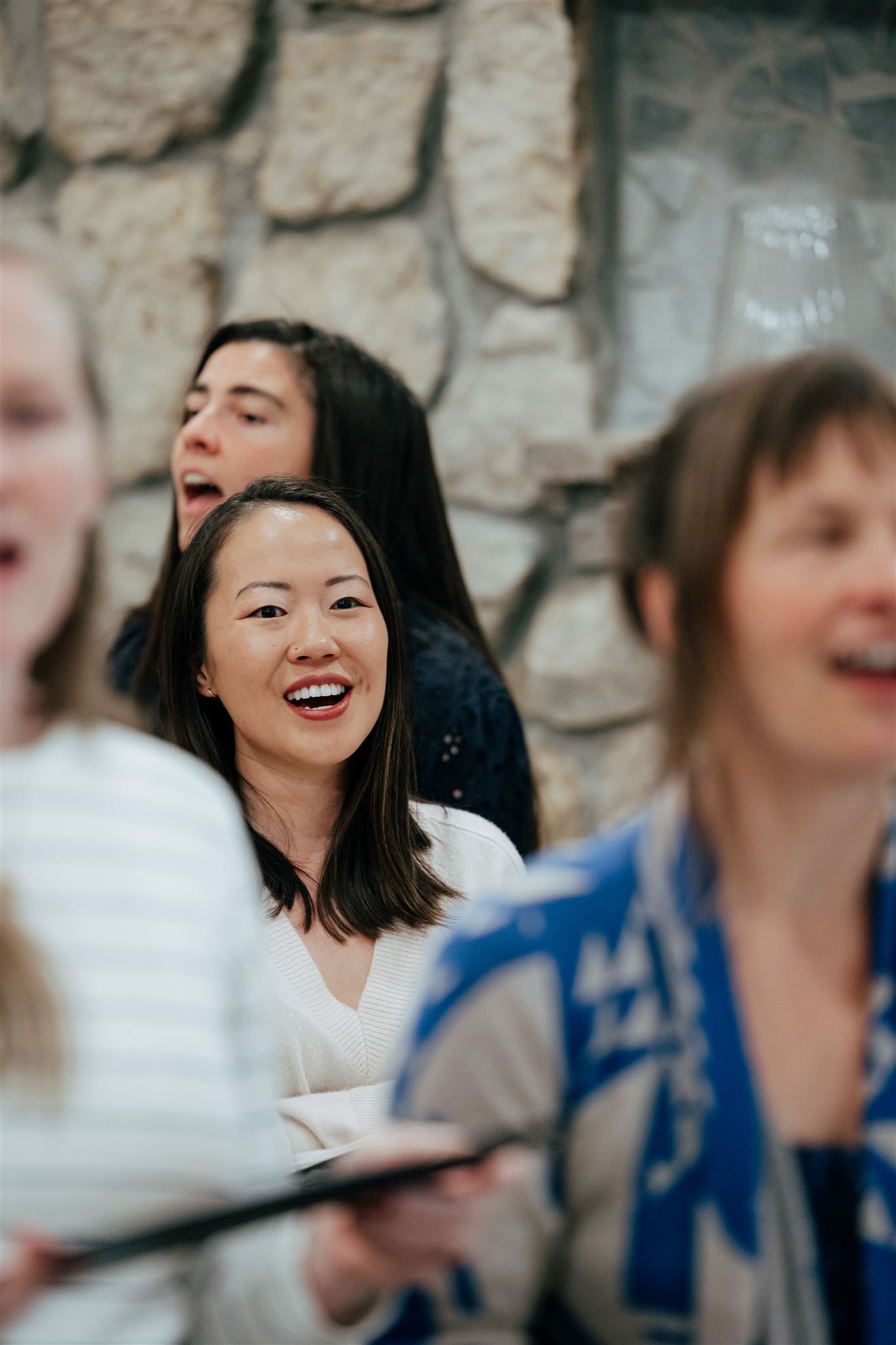 Group of people singing in a choir, with a stone wall in the background.