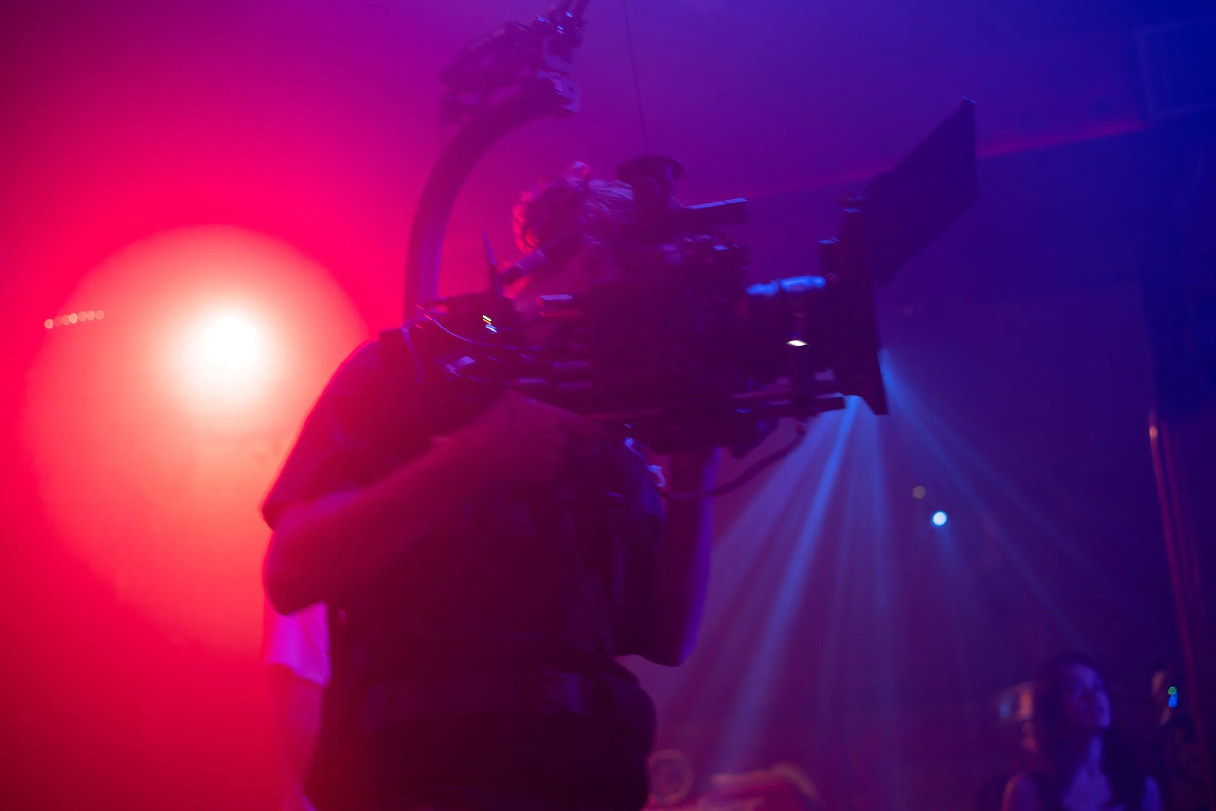 A person filming with a professional camera in a dimly lit room with red and blue stage lighting.