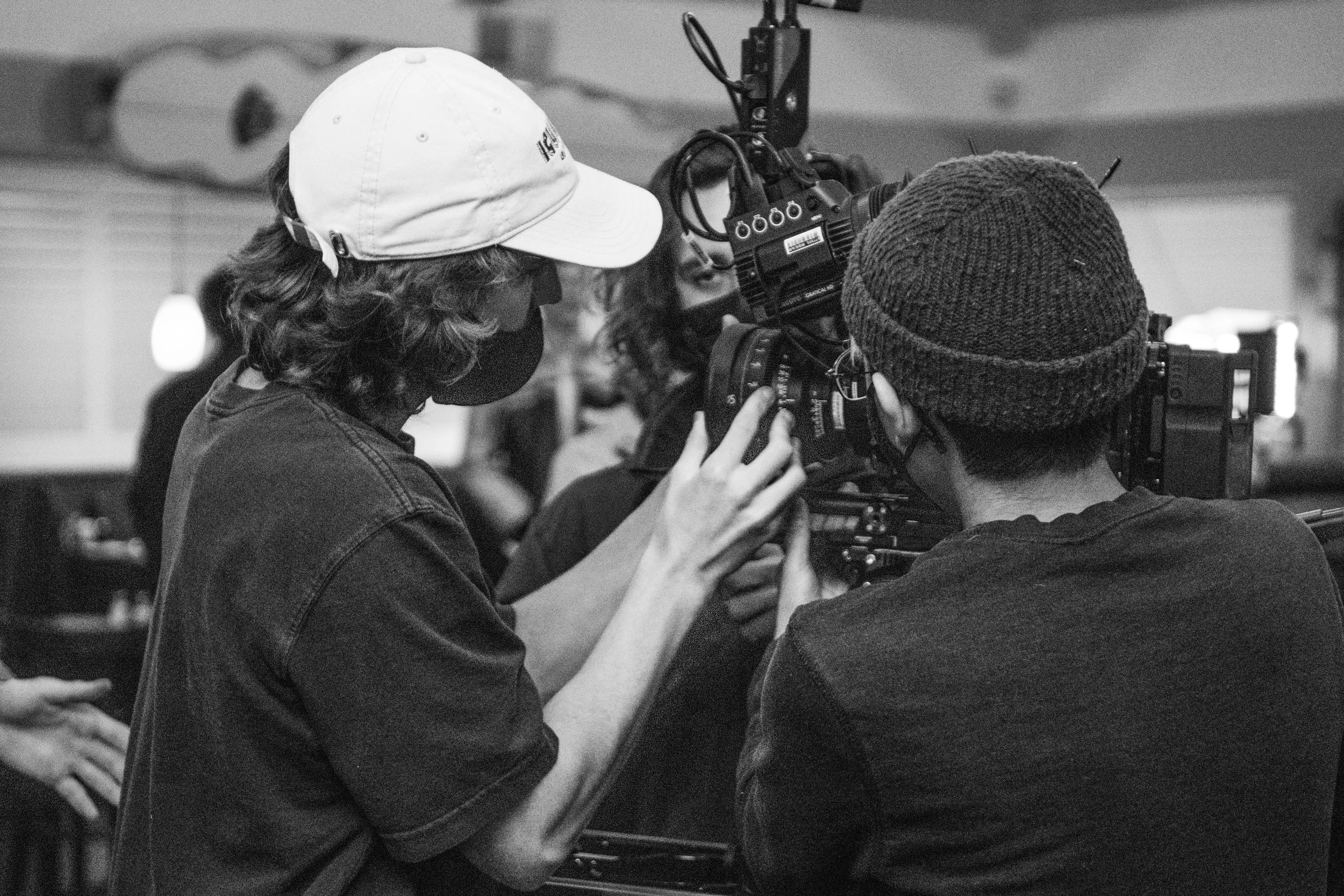 Two people operating a professional video camera, one wearing a baseball cap and mask, the other wearing a beanie, in a behind-the-scenes setting.
