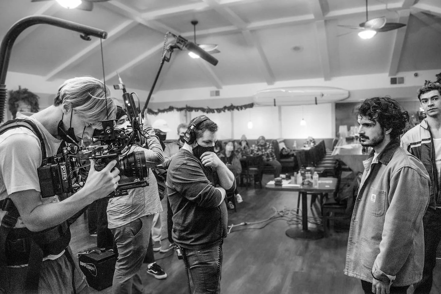Black and white photo of a film set showing a camera operator filming a scene with two actors, one with curly hair and a jacket, and another with a face mask, in a room with people seated in background.