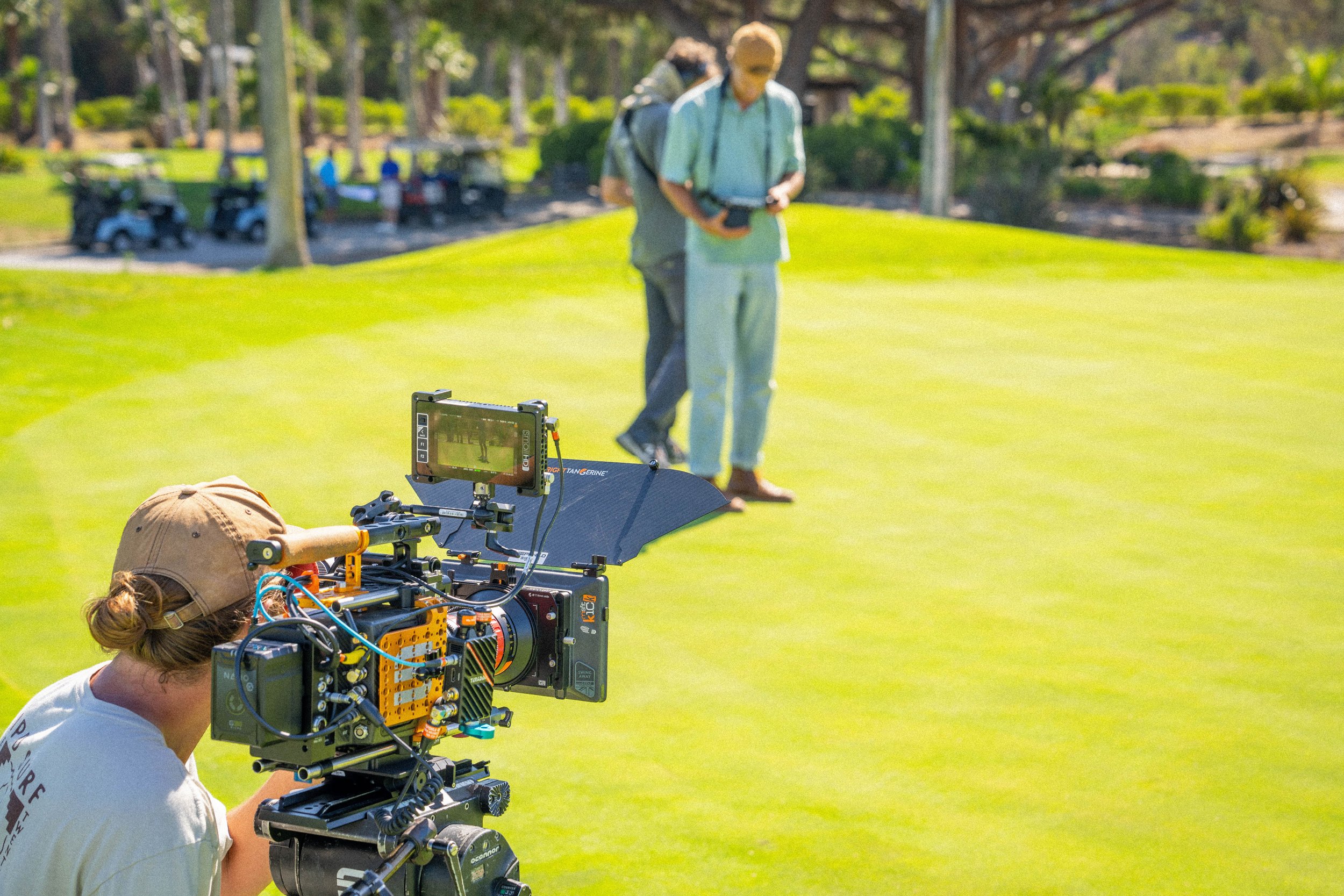 A film crew setting up a shot on a golf course with two people in the background, one filming and the other preparing to play, while the camera team prepares the camera.