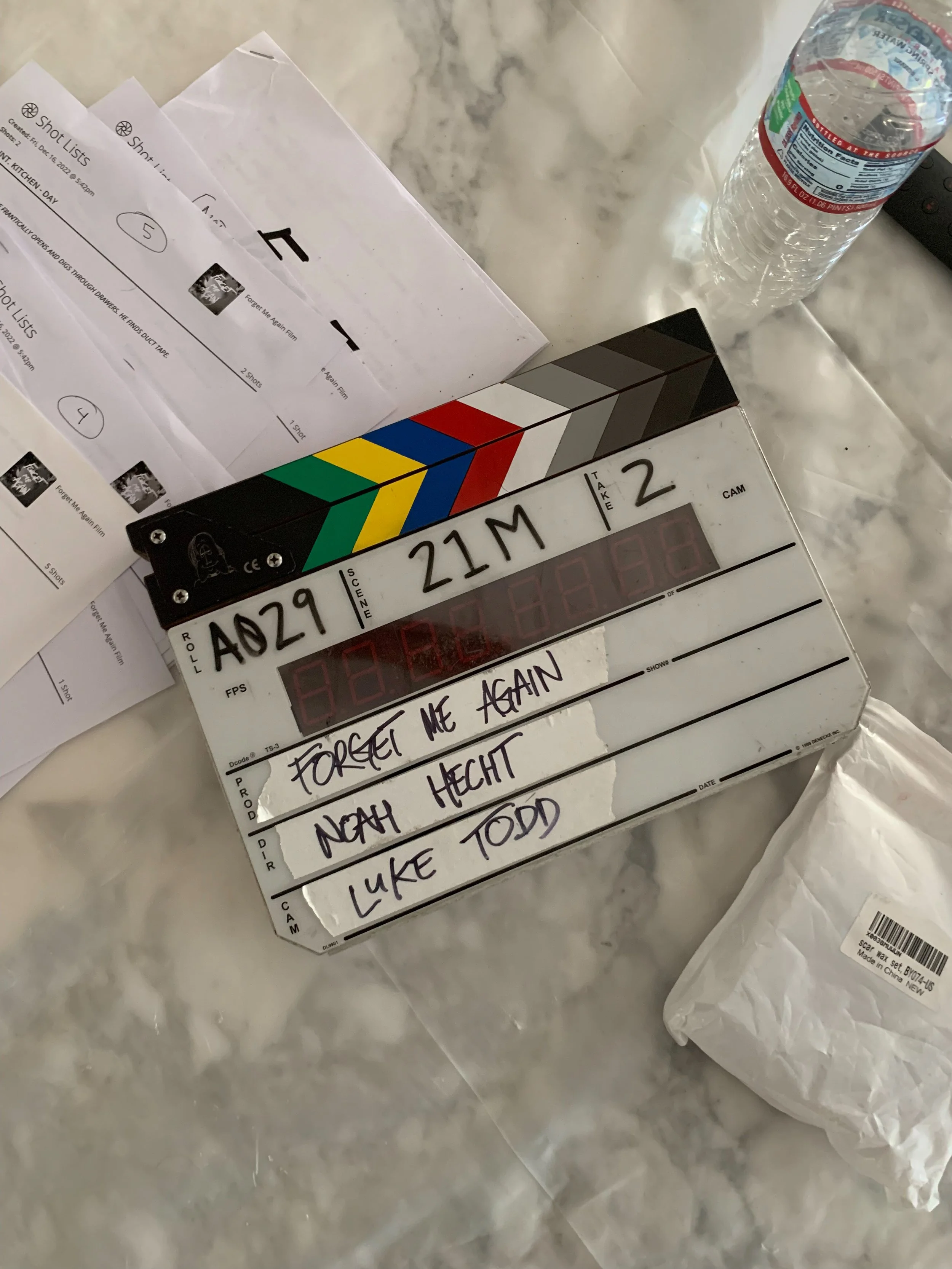 Clapperboard, notes with the names 'Forget Me Again', 'Noah Hecht', and 'Luke Todd', a water bottle, and some papers on a marble surface.