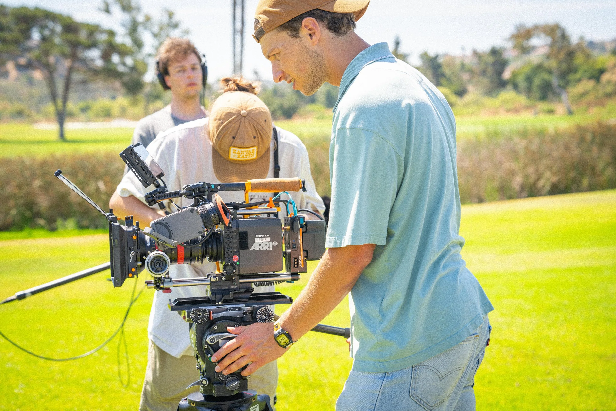 Filmmakers on a movie set outdoors, with a camera on a tripod in focus, and crew members adjusting equipment, in a grassy area with trees and blue sky.