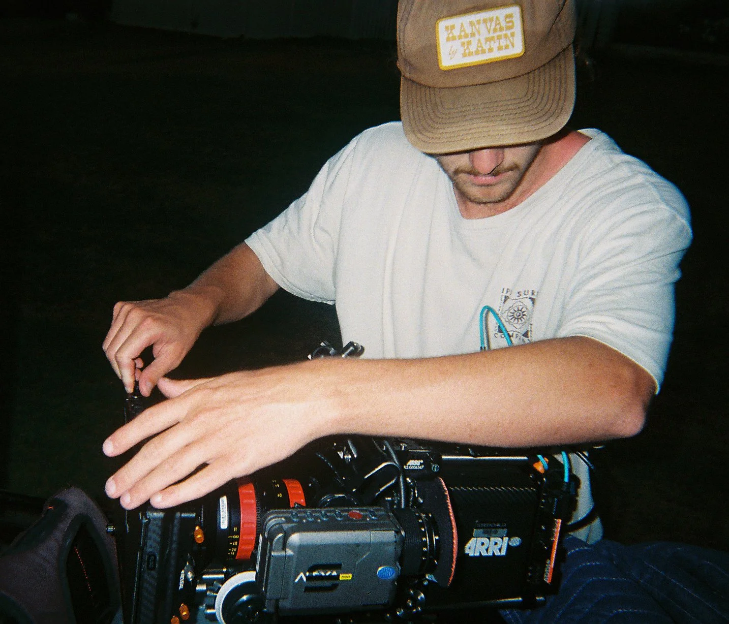 A man with a beige cap and white t-shirt working with a professional video camera outdoors at night.