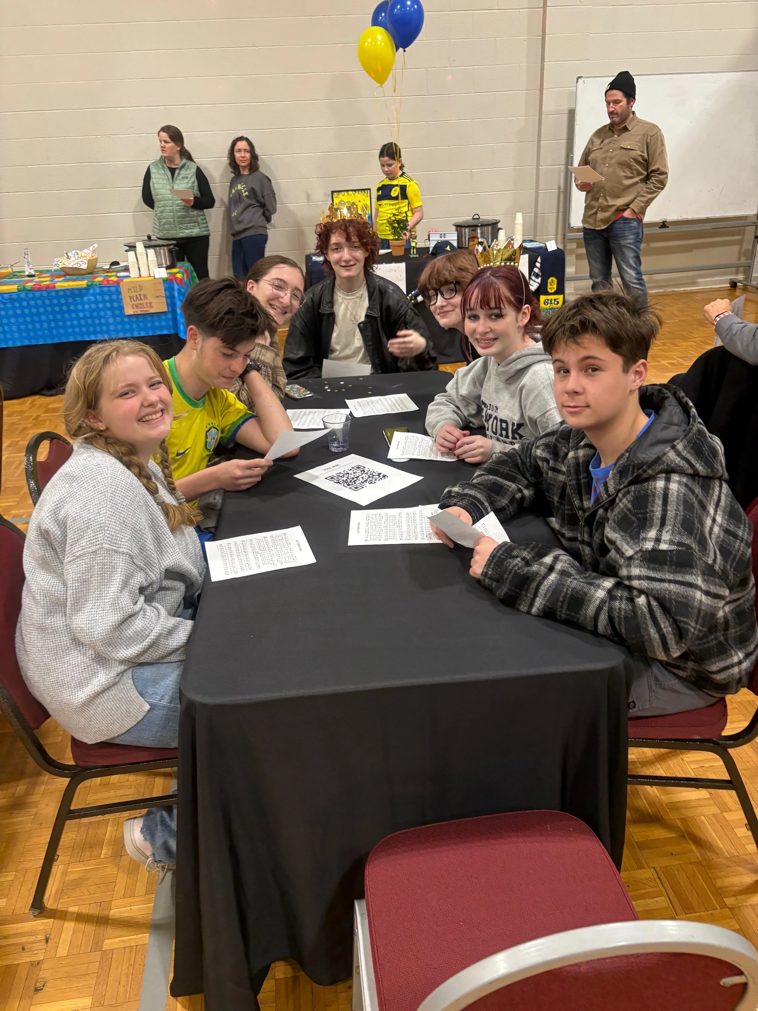 A group of children sitting around a black table, smiling and holding papers, at a party or event. In the background, there are balloons, a table with snacks, and a few adults standing near a whiteboard.