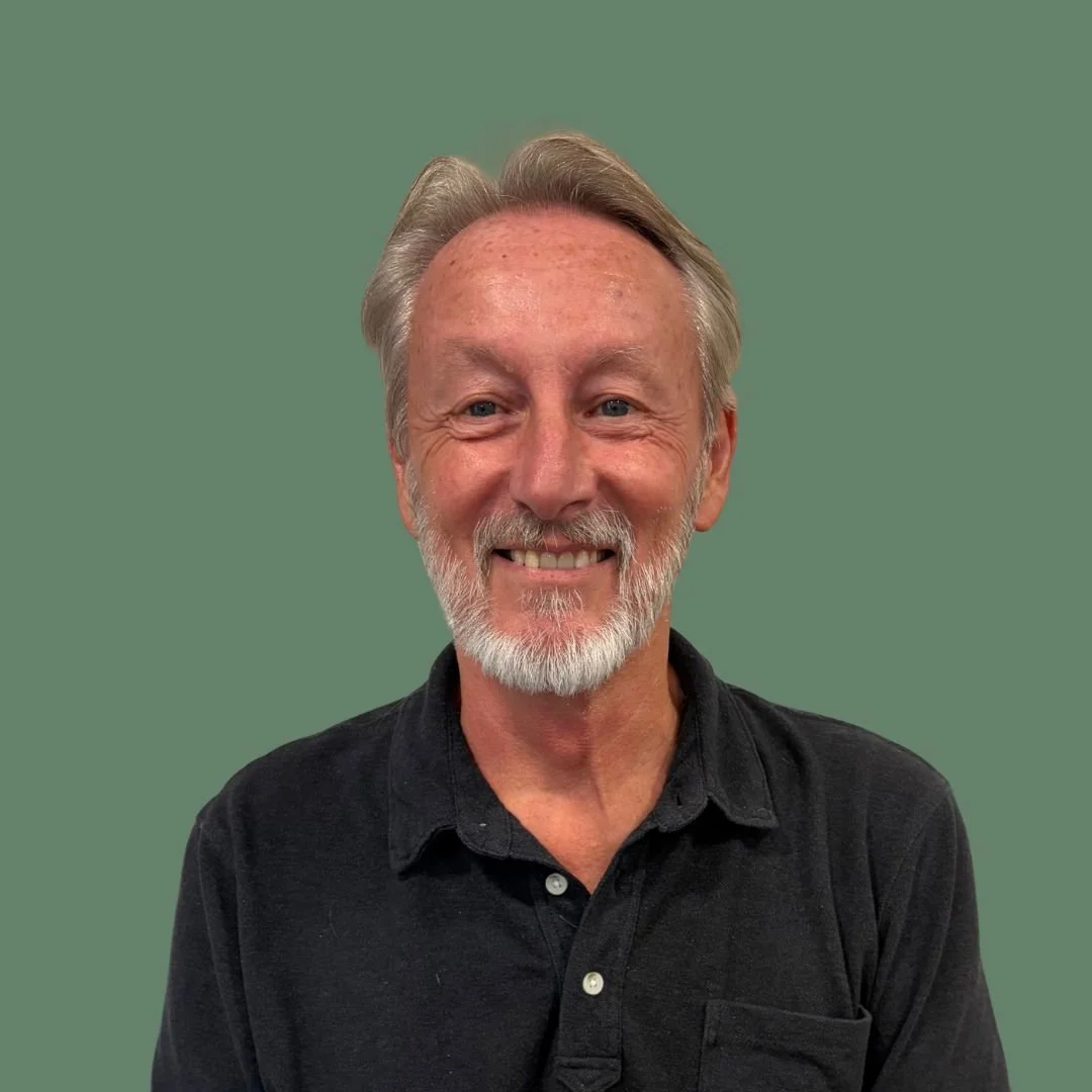 Portrait of a smiling man with gray hair and a beard, wearing a black shirt, against a green background.