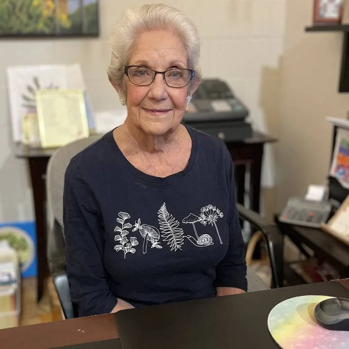 An elderly woman with glasses and white hair, sitting at a desk in an office, wearing a navy shirt with white plant illustrations.