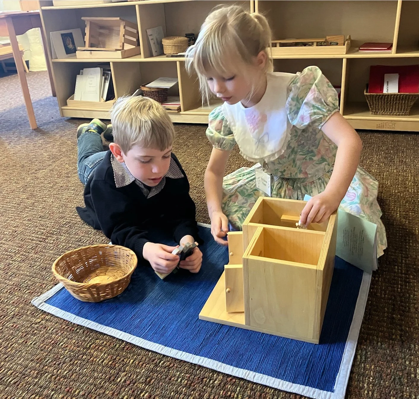 A young boy and girl lying on a blue rug in a classroom, engaging in a pretend play with wooden toys and a wicker basket.