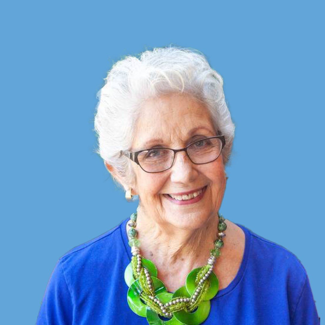 An elderly woman with white hair and glasses smiling, wearing a blue shirt and a large green beaded necklace against a light blue background.