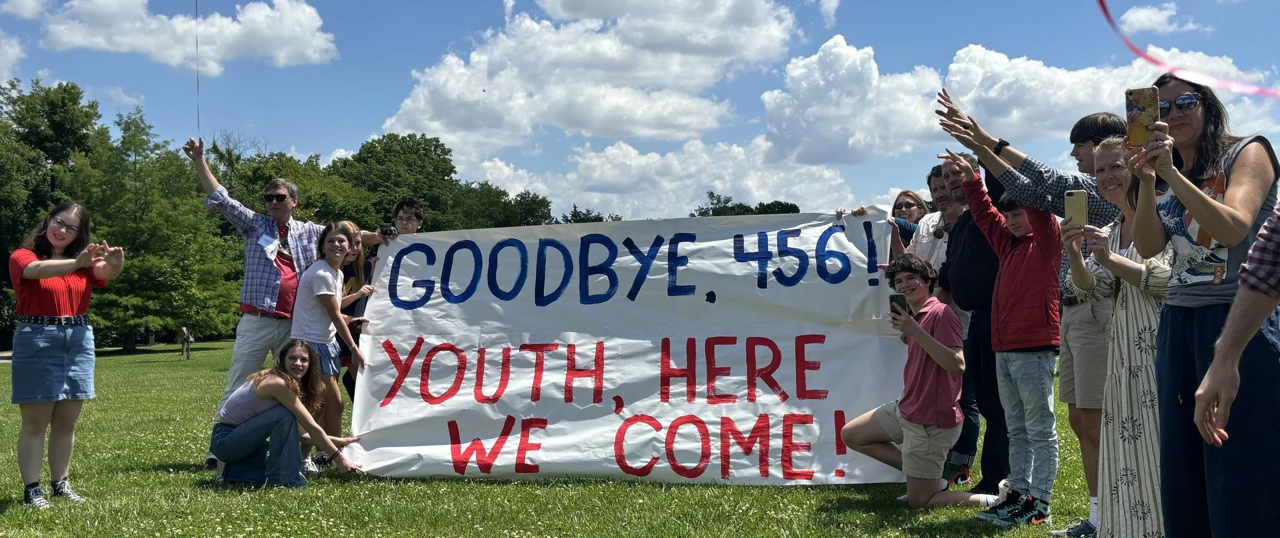 Group of people outdoors holding a large banner that reads, 'Goodbye, 456! Youth, here we come!' Some are taking photos and others are smiling and posing.