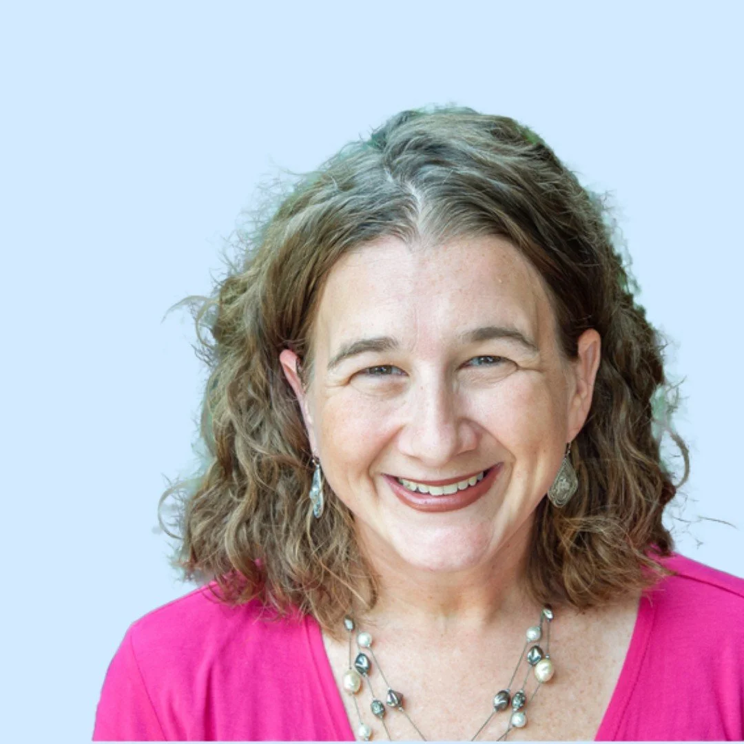 A woman smiling, wearing a pink top, earrings, and a necklace, with curly shoulder-length brown hair, against a light blue background.