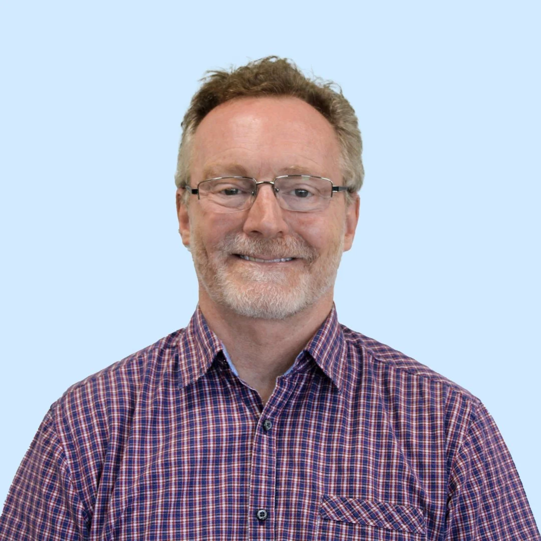 A middle-aged man with glasses, light brown hair, and a beard, smiling, wearing a checked shirt, standing against a light blue background.