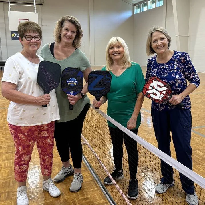 Four women holding pickleball paddles standing on an indoor pickleball court with a net.