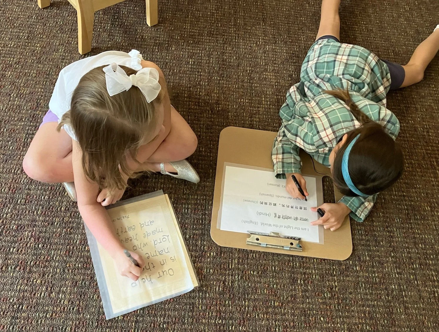 Two young girls lying on the carpeted floor, one with a large white bow in her hair, working on writing assignments with paper and markers. One girl is on a small clipboard with print, the other on a laminated sheet with handwriting, placed next to a