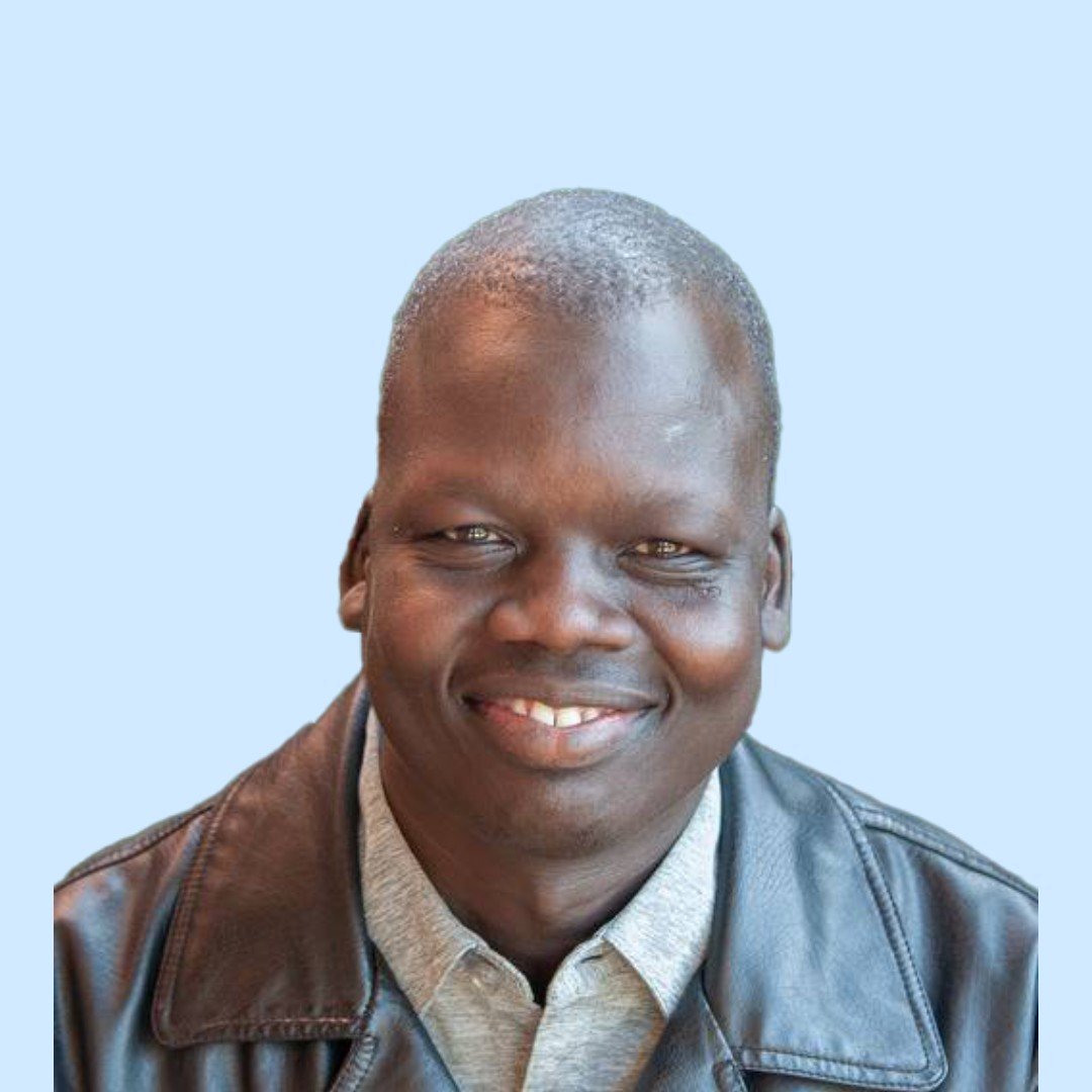 A smiling man with short hair wearing a leather jacket and light-colored shirt, against a light blue background.