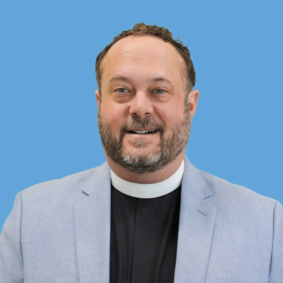 A man with a beard wearing a clerical collar, black shirt, and light-colored blazer, smiling against a blue background.