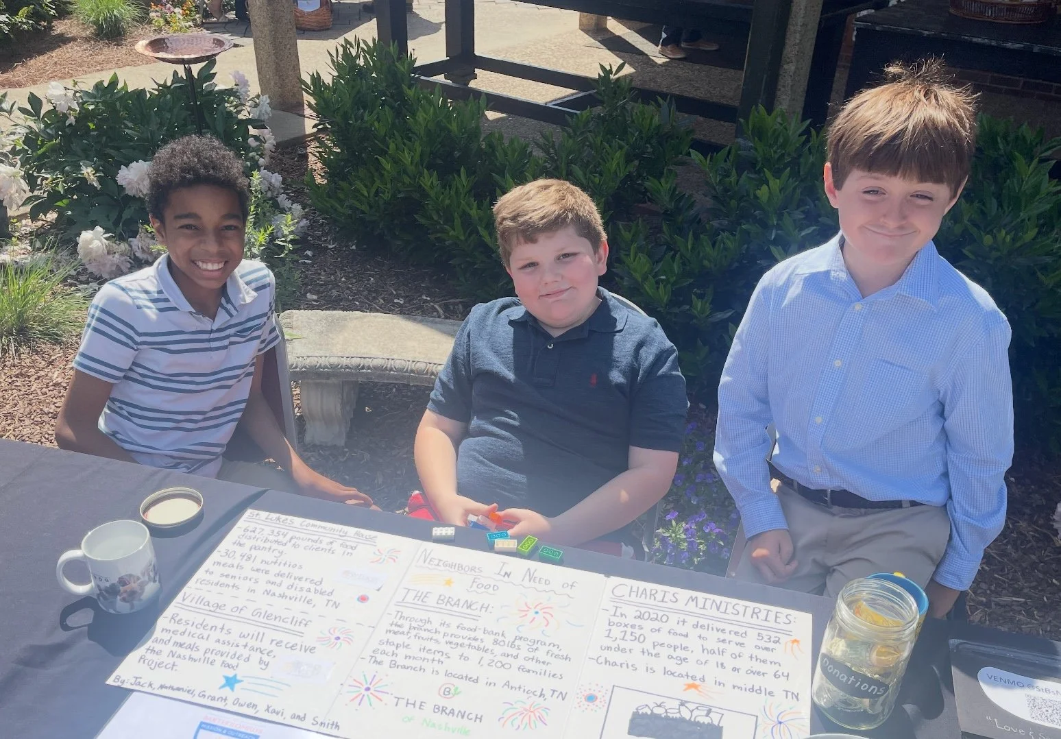 Three boys sitting outdoors at a table with informational posters, books, and a jar of donations visible, smiling in the sunlight.