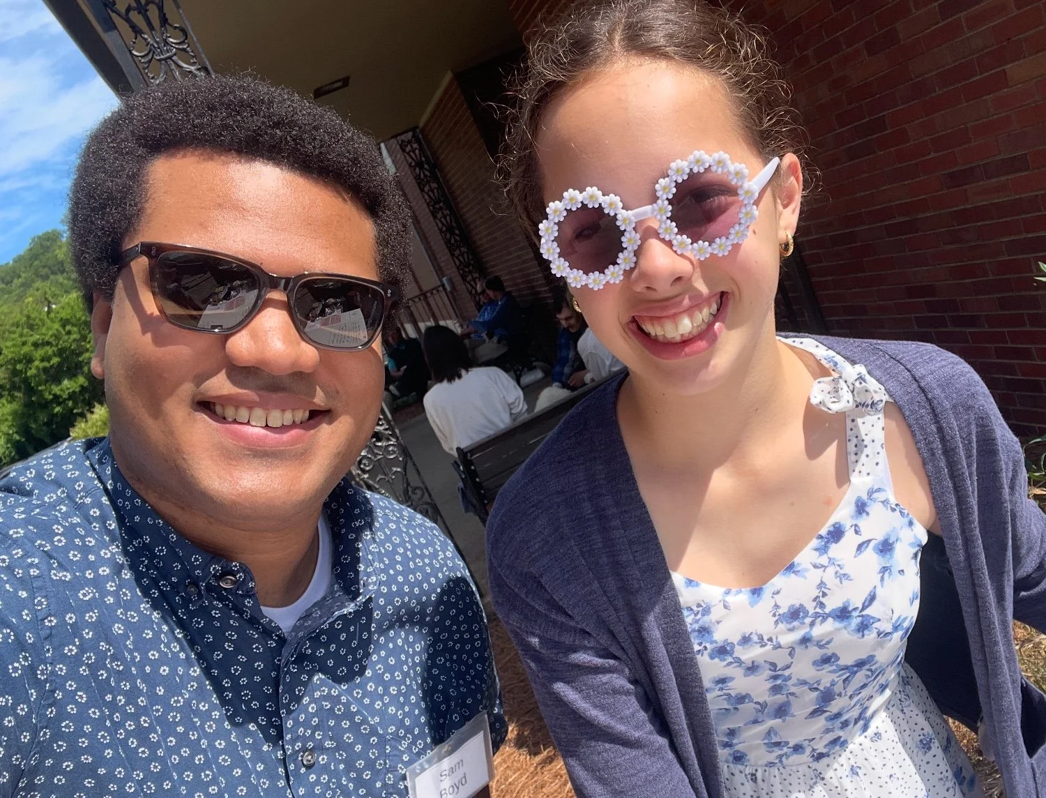 A man and a woman taking a selfie outdoors on a sunny day. Both are smiling. The man is wearing sunglasses and a blue patterned shirt. The woman is wearing flower-shaped sunglasses with daisies and a floral dress with a cardigan.