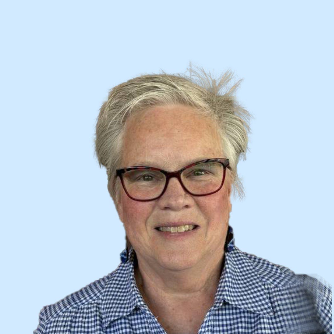 A woman with short gray hair, glasses, and a checkered shirt smiling at the camera against a light blue background.
