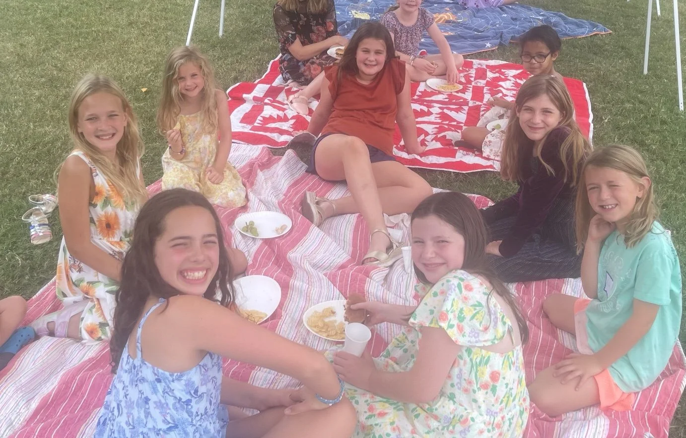 Group of children having a picnic outdoors on a grassy area, sitting on blankets with plates of food.