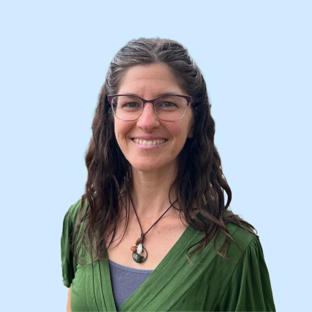 A woman with glasses, smiling, wearing a green top and a necklace, standing in front of a blue background.