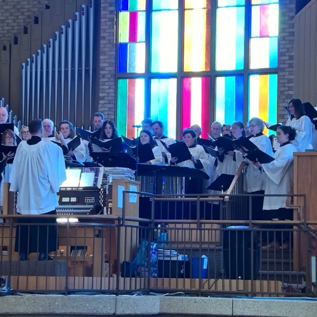 A choir of people dressed in white robes performing in a church. The background features a large colorful stained glass window.