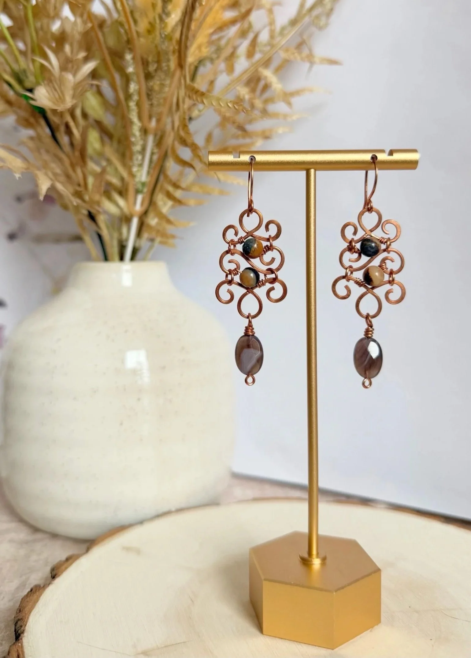 A pair of copper earrings with decorative loops and beads, hanging from a gold-colored stand on a beige surface, with dried ornamental grass in a white vase in the background.