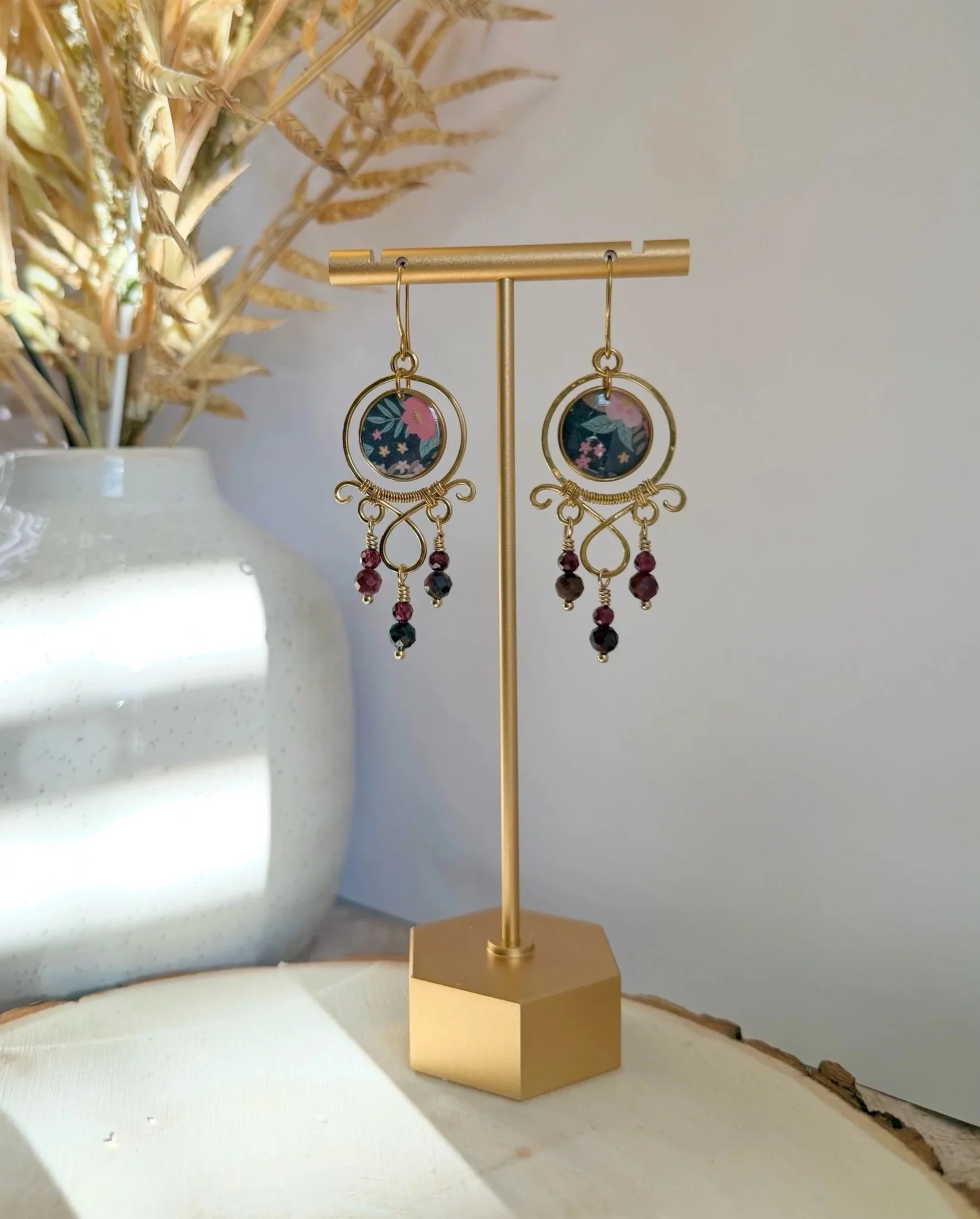 Gold jewelry display stand with circular earrings on a beige pedestal, next to a white ceramic pot with dried yellowish foliage.