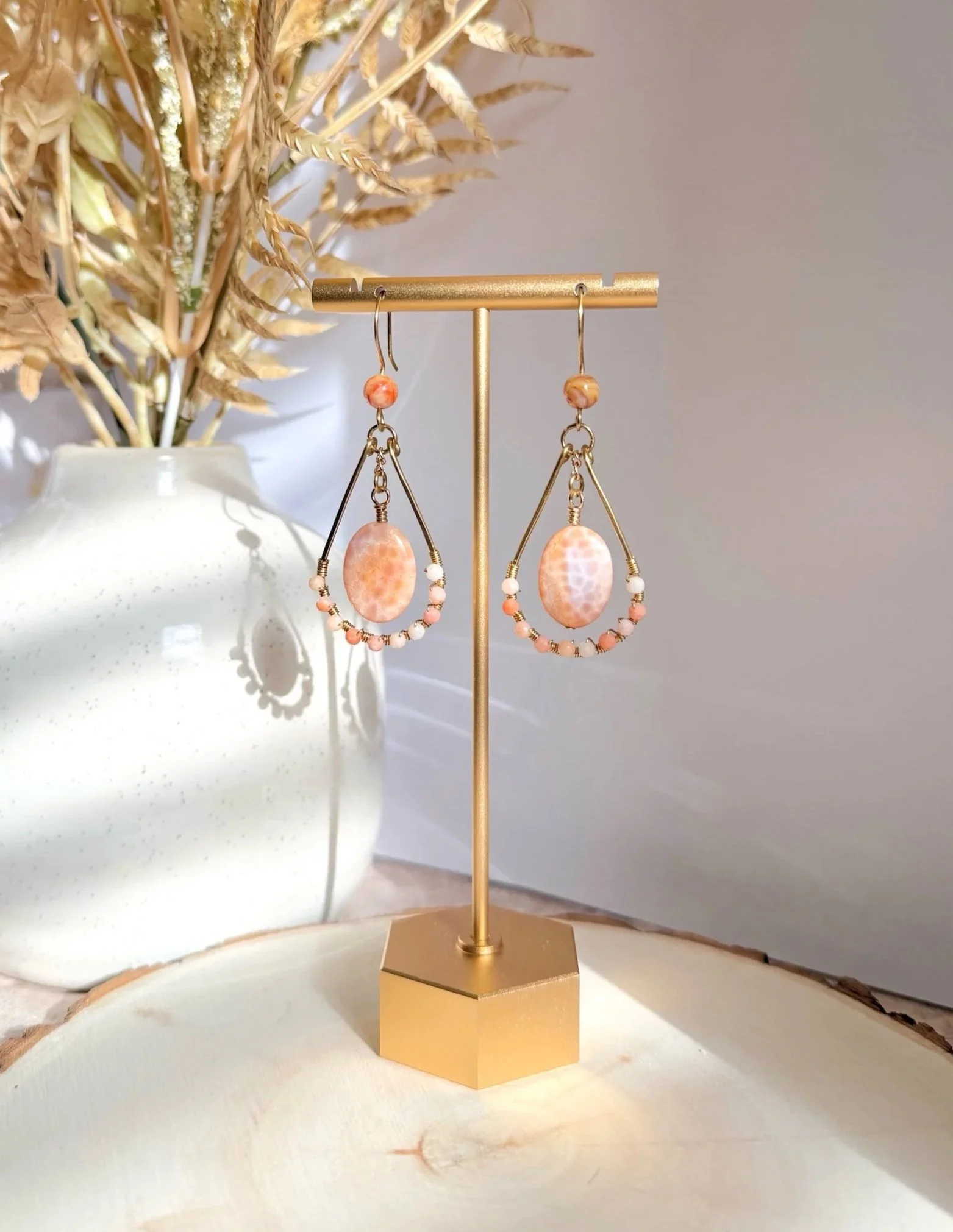 Gold jewelry display stand with a pair of peach-colored drop earrings and a matching bracelet, placed on a white surface with a dried floral arrangement in a white vase in the background.