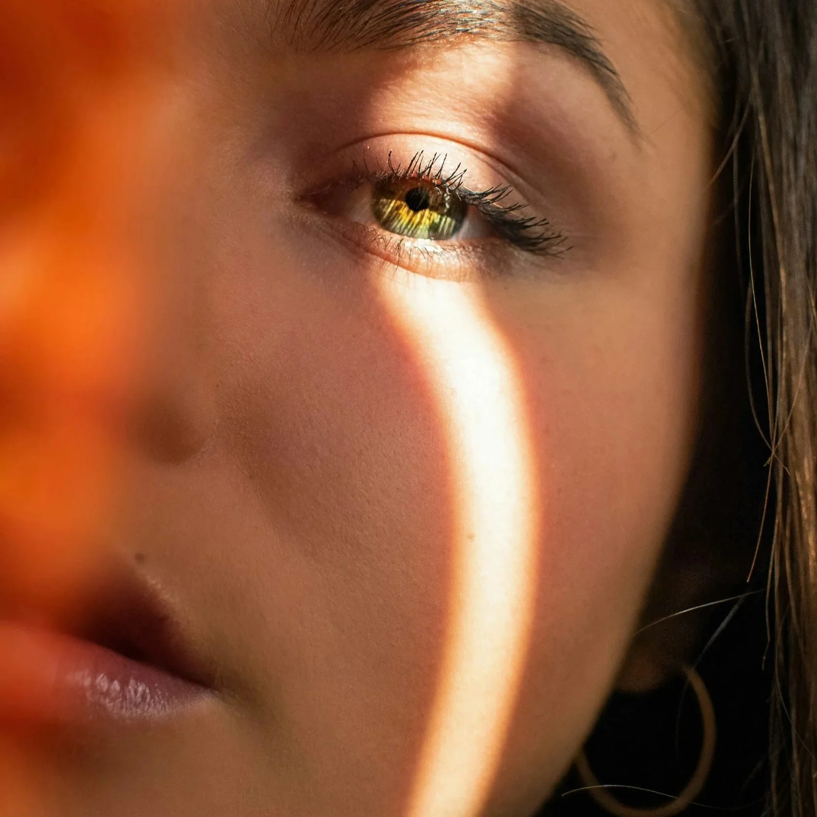 Close-up of a woman's face with a narrow band of sunlight cast across her nose and cheek, emphasizing her eye with a reflection of outdoors in the iris.
