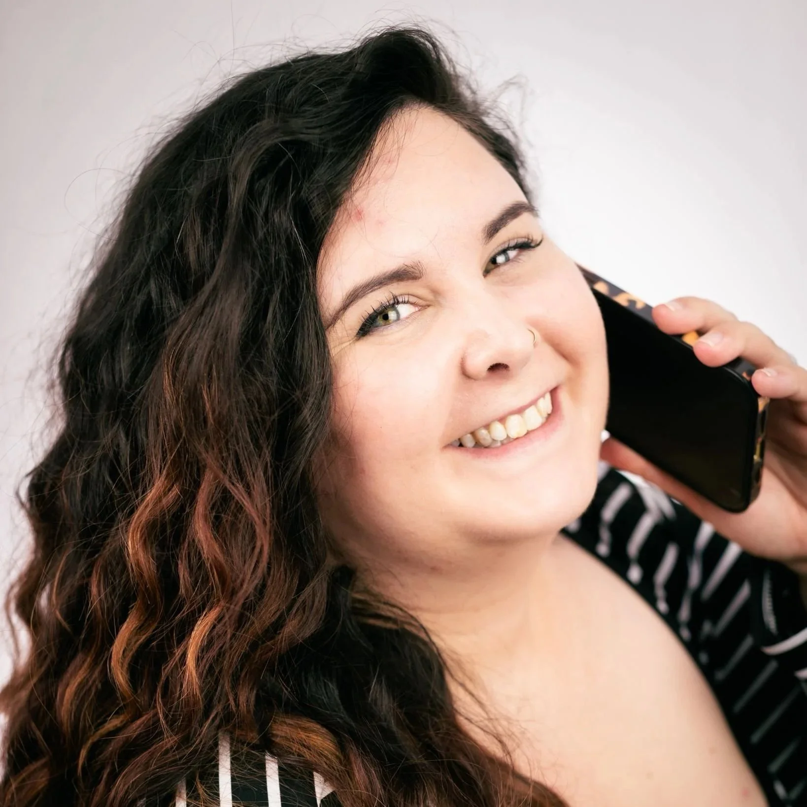 A woman with long, wavy dark hair with reddish-brown tips is smiling and speaking on a cell phone.