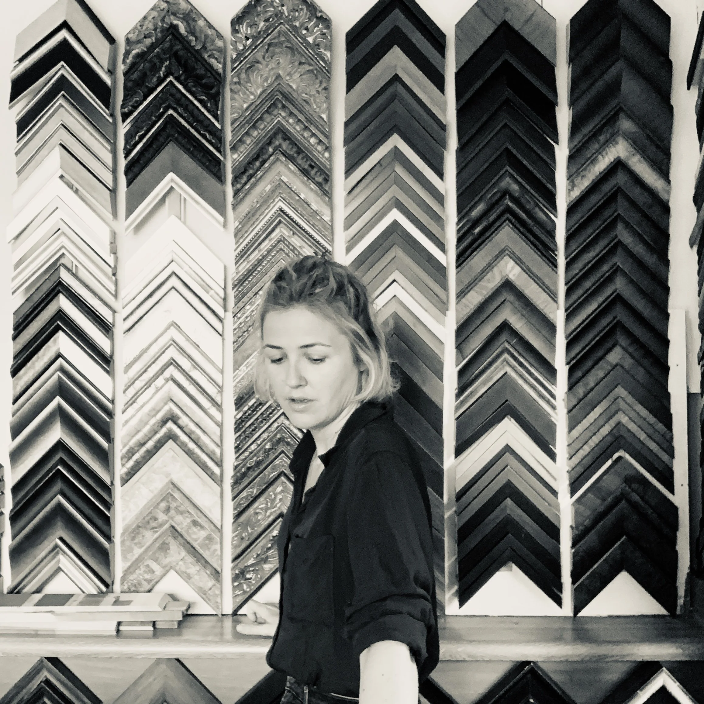 A woman with light hair styled in a bun with loose strands, wearing a black blazer, standing in front of a display of various black, white, and patterned picture frames arranged vertically. She is looking downward.