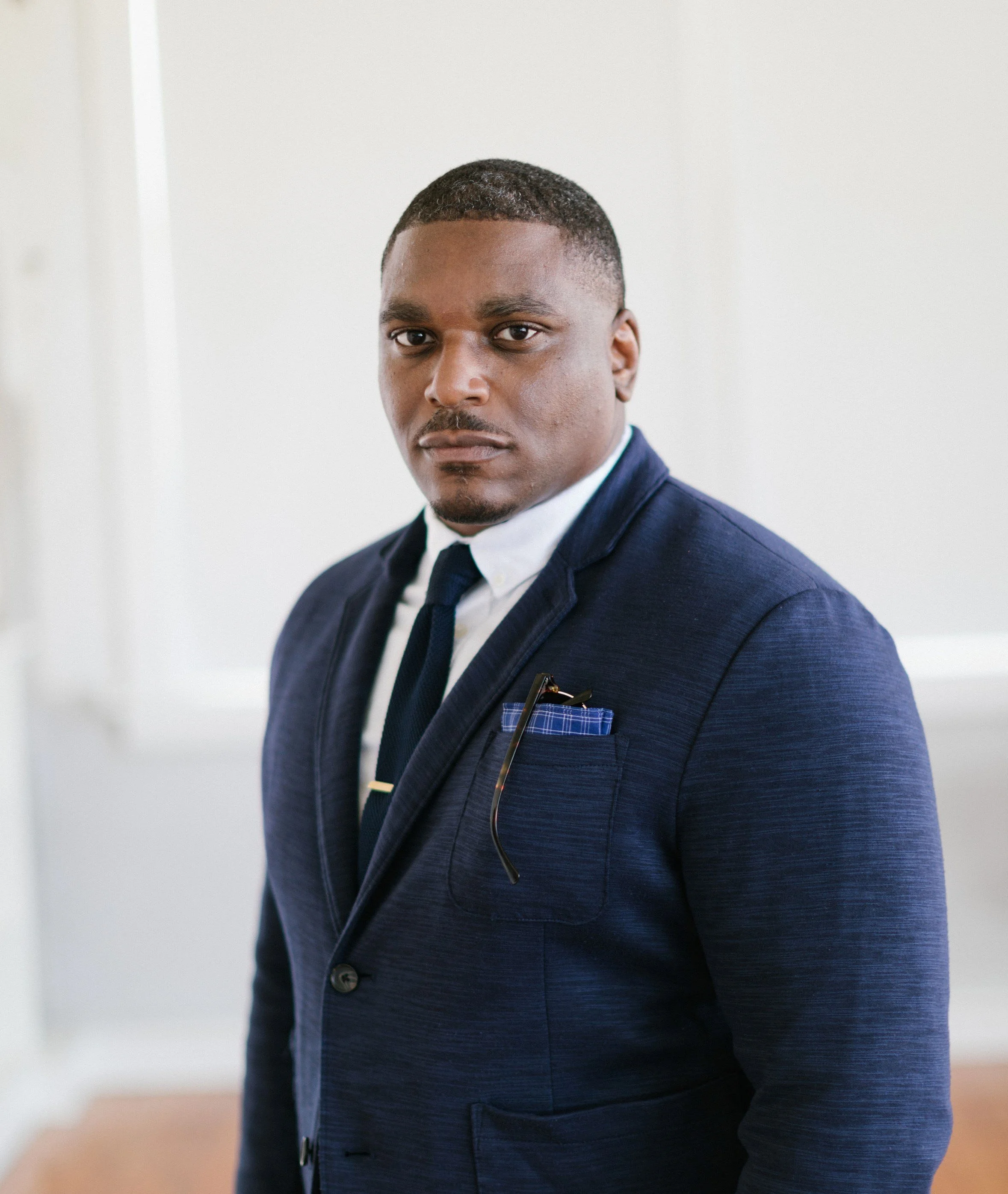 A man wearing a navy blue suit, white shirt, and navy tie, standing indoors against a white wall, looking directly at the camera with a serious expression.