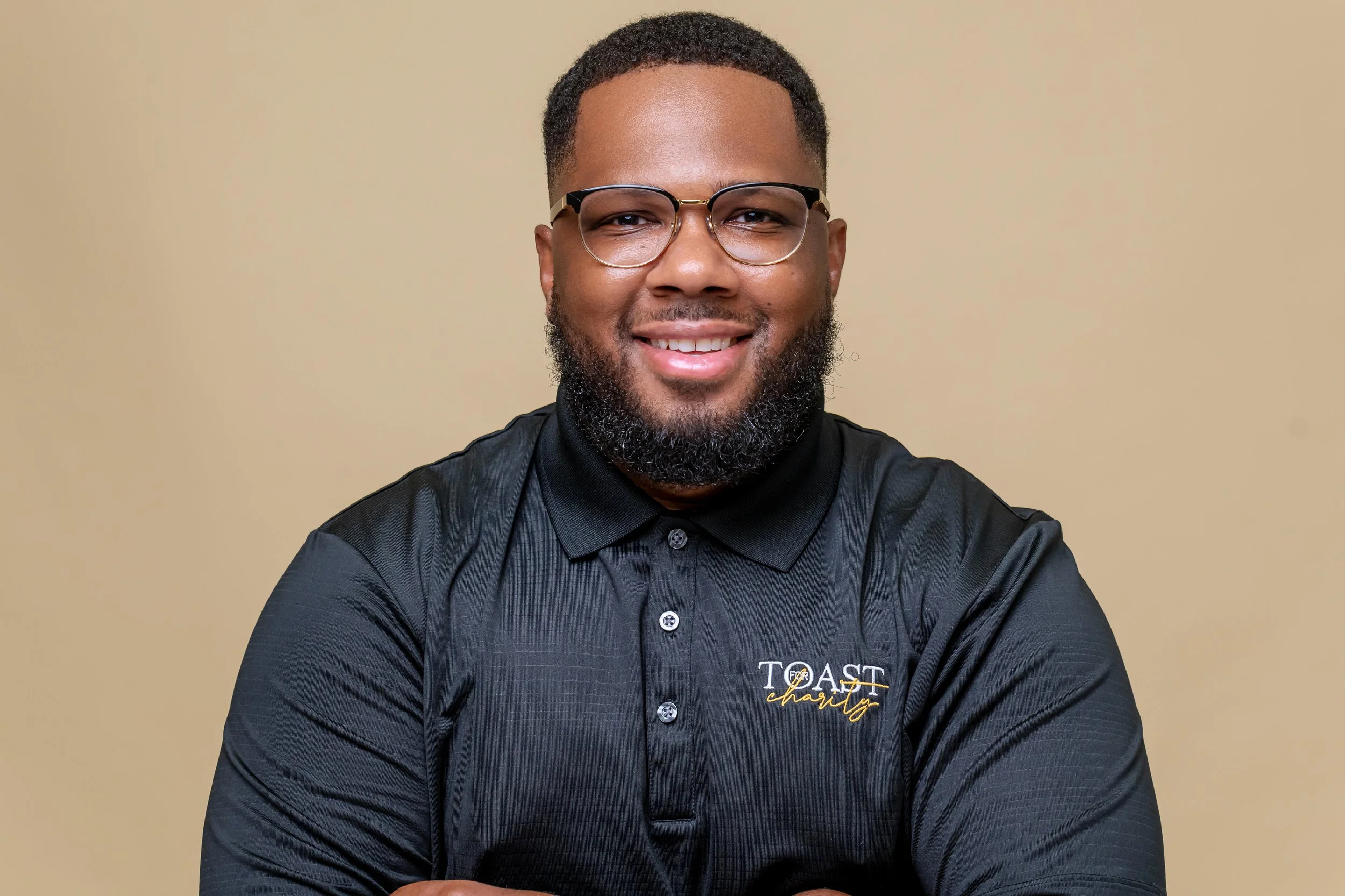 A smiling man with glasses and a beard, wearing a black polo shirt with the logo 'TOAST charity' on it, standing against a beige background.