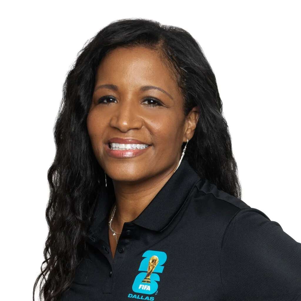 A woman with long, curly black hair is smiling and wearing a black collared shirt with a FIFA Dallas logo.