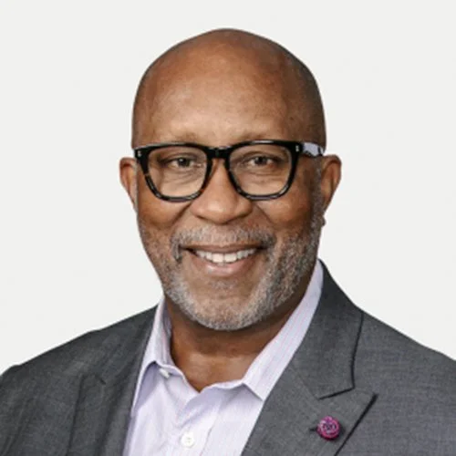 A middle-aged African American man with a shaved head, wearing black framed glasses, a gray suit jacket, and a light purple dress shirt, smiling.