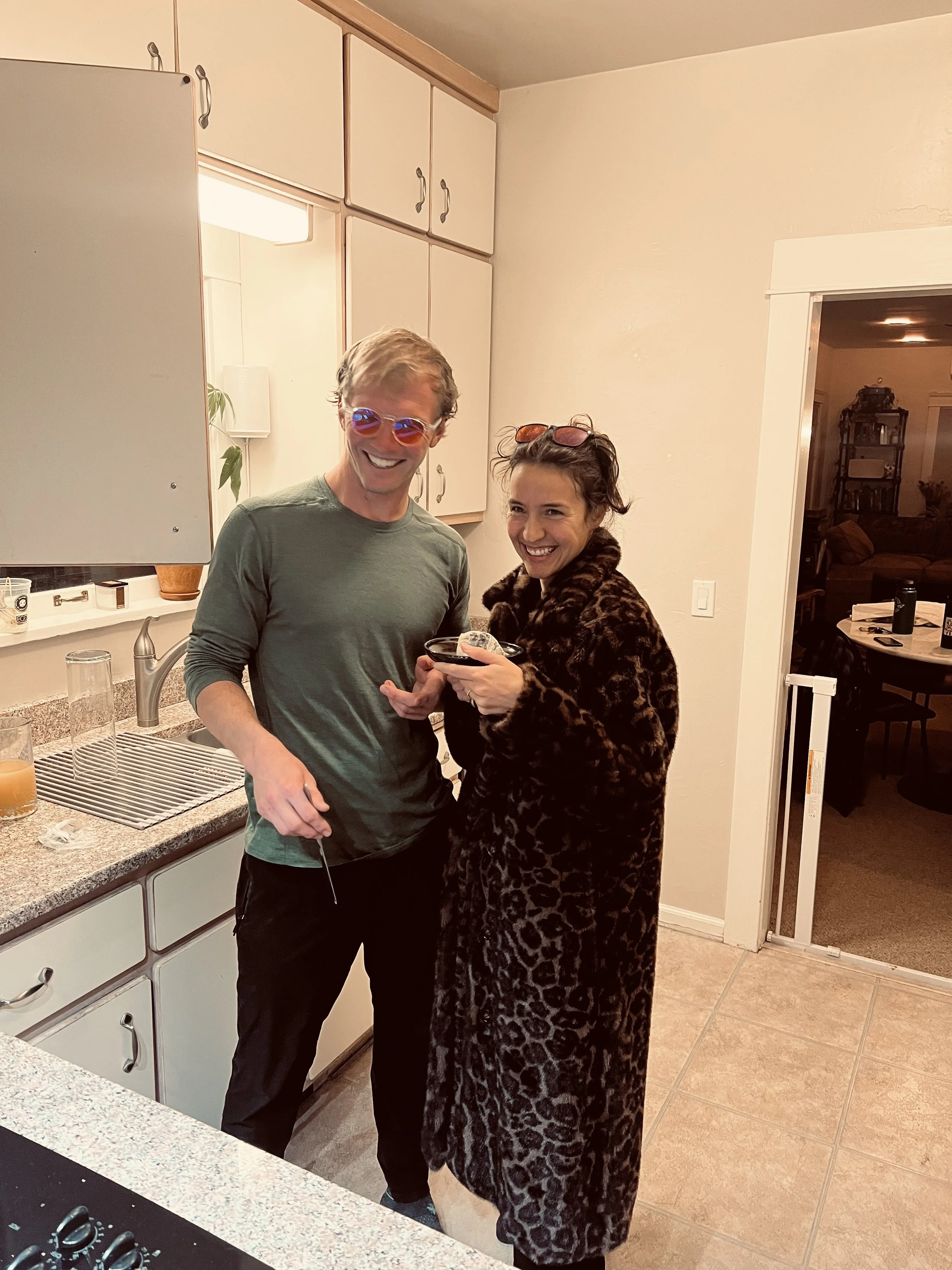 Two people smiling and looking at the camera in a kitchen, wearing sunglasses. The person on the left is a man with light hair and a green shirt, holding a drink, and the person on the right is a woman with dark hair wearing a leopard print coat. There are cups and glasses on the kitchen counter.